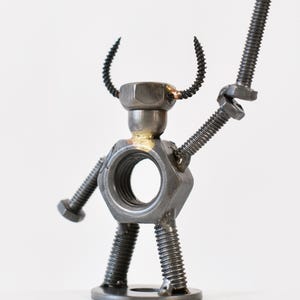 May include: A handcrafted metal sculpture of a humanoid figure, constructed from nuts, bolts, and screws. The figure has a hexagonal nut for a torso, a bolt held aloft as a weapon, and curved wire horns. The sculpture is on a white background.