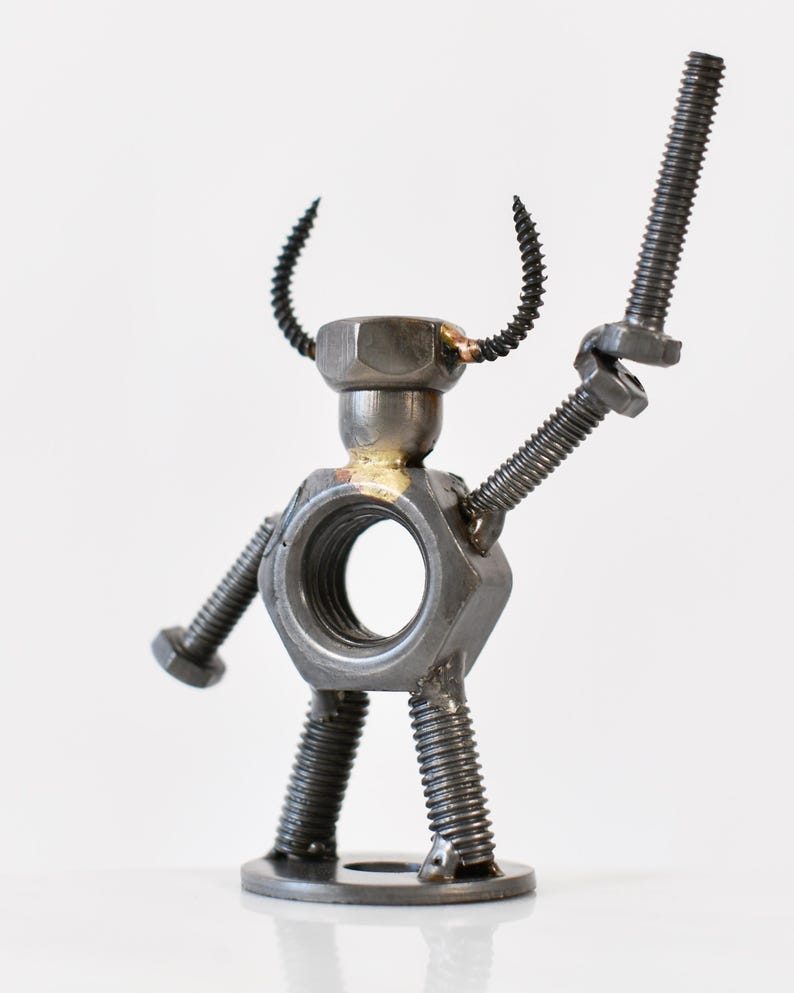 May include: A handcrafted metal sculpture of a humanoid figure, constructed from nuts, bolts, and screws. The figure has a hexagonal nut for a torso, a bolt held aloft as a weapon, and curved wire horns. The sculpture is on a white background.