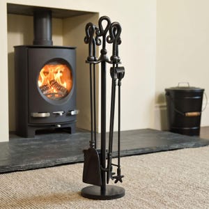 May include: A black wrought iron fireplace tool set featuring a stand with a circular base and three decorative looped handles. The set includes a shovel, a poker, and a brush, all suspended from the central vertical support rod.