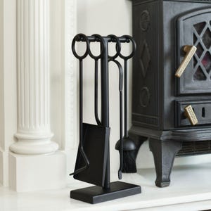 May include: A black fireplace tool set with a stand. The set includes a poker, shovel, brush, and a tool with a curved end. The tools have ring handles and are displayed in front of a black stove.