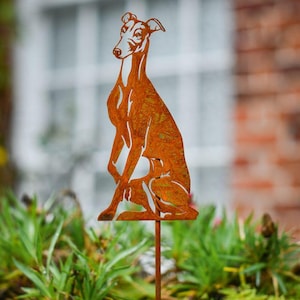 May include: A weathered, rusty brown metal garden ornament depicting a seated greyhound dog. The dog's form is cut out of the metal and mounted on a thin stake. The ornament is placed in a garden bed with green foliage.