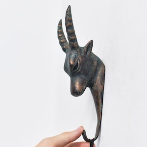May include: A bronze-effect wall hook in the shape of an antelope head. The hook is fixed to a white wall, with the antelope's head and curved horns pointing upwards. A hand is holding a black item on the hook.