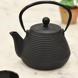 May include: A black cast iron teapot with a curved handle and a small knob on the lid. The teapot has a textured, ribbed design and a spout for pouring. The teapot is designed for brewing tea.
