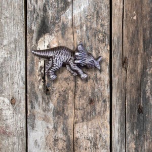 May include: A grey metal dinosaur figurine with a textured surface. The dinosaur is in a running pose with its mouth open. The figurine is on a wooden background.