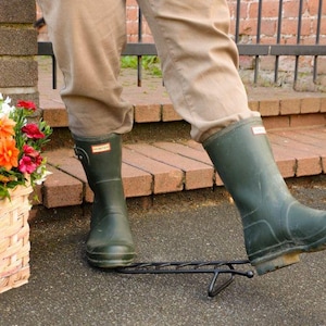 May include: A black metal boot jack with a U-shaped opening for removing footwear, positioned on the ground. The device features a sturdy frame with a heel rest and a base plate, shown in use with dark olive green rubber boots and tan trousers.