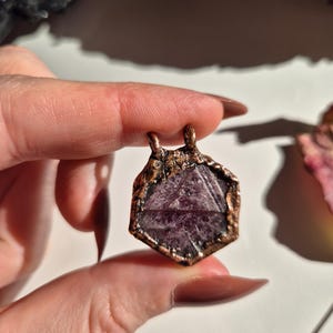 May include: A hexagonal pendant featuring a deep purple, faceted gemstone encased in a textured, dark copper-toned metal frame. The pendant includes two small metal loops at the top for attachment, showcasing a raw, metallic finish throughout.