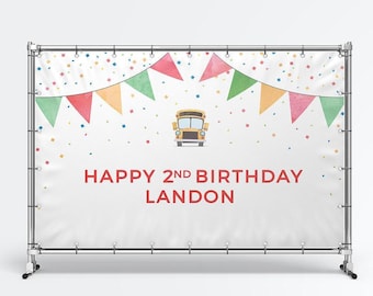 May include: A white rectangular birthday backdrop banner mounted on a silver metal frame. The design features colorful triangular bunting flags, scattered confetti, a yellow school bus illustration, and the text HAPPY 2ND BIRTHDAY LANDON.