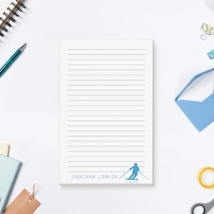 May include: A white notepad with blue lines and a blue silhouette of a skier at the bottom. The notepad has the name "Jonathan Lawlor" printed at the bottom.