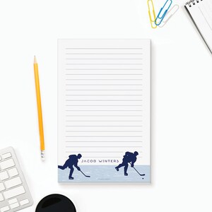 May include: A lined notepad featuring a blue silhouette of hockey players. The notepad is personalised with the name "Jacob Winters".