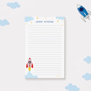 May include: A personalized notepad with a rocket ship design. The notepad features the name "JAYDEN PETERSON" in blue, with stars and clouds. The rocket ship is red, white, and yellow. The notepad has lined pages.