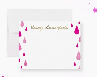May include: A white stationery set featuring a notepad and envelope. The notepad has a gold script name and pink watercolor raindrop designs. Pink confetti and stars are scattered around the set.