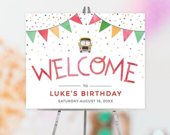 May include: A rectangular birthday sign on a metal easel featuring a yellow school bus icon, colorful bunting, and confetti. The text reads: WELCOME TO LUKE'S BIRTHDAY SATURDAY AUGUST 18, 20XX. The design uses red, green, and orange accents.