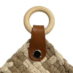 May include: A beige and white woven wall hanging with a brown leather loop and a wooden ring. The loop has a metal button.