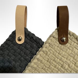 May include: Two woven kitchen pot holders with leather hanging loops. One pot holder is dark gray and the other is beige. Both have a silver metal button on the loop.