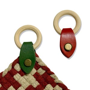 May include: Two woven pot holders with wooden rings and leather accents. One pot holder has a green leather accent, and the other has a red leather accent. The pot holders are red and beige in a checkered pattern.