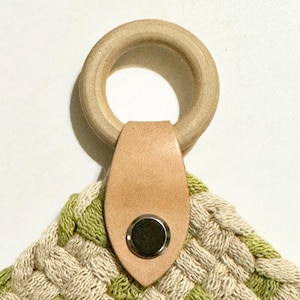 May include: A woven wall hanging with a green and white pattern. The hanging has a wooden ring and a leather strap with a metal button.
