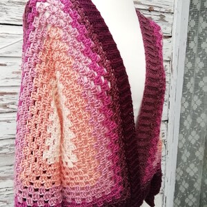 Può includere: Un cardigan all'uncinetto con scollo a V e maniche lunghe. Il cardigan è realizzato in un gradiente di filato rosa, pesca e bordeaux.