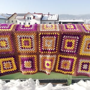 Könnte beinhalten: Handgefertigte Häkeldecke mit Patchwork-Design. Die Decke zeigt eine lebendige Farbpalette aus gelben, lila und braunen Quadraten. Die Decke wird im Freien über einer grünen Oberfläche drapiert, mit einem verschneiten Hintergrund.