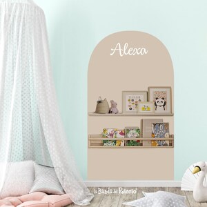Op de afbeelding: Een muursticker in de vorm van een beige boog met de naam Alexa in wit handschrift, twee horizontale planken met diverse boeken en kleine decoratieve items, een witte hemel, een roze vloerkussen en een stervormig kussen.