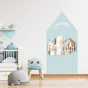 Könnte beinhalten: Eine hellblaue Wand mit einem weißen, hausförmigen Aufkleber, auf dem "Charlotte" steht. Der Aufkleber hat eine Reihe kleiner weißer Regale im Inneren. Die Regale sind mit kleinen Holzspielzeugen und Büchern gefüllt. Im Vordergrund befinden sich ein weißes Babybett, ein graues Wal-Plüschtier und ein blauer Stuhl mit einem wolkenförmigen Sitz.