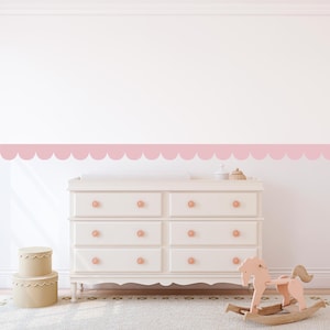 Può includere: Una cassettiera bianca con sei cassetti e pomelli rosa, un cavallo a dondolo rosa e due scatole rotonde beige. Un bordo rosa smerlato adorna la parete sopra la cassettiera. La stanza ha un'estetica morbida e invitante.