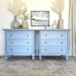 May include: Two light blue bedside tables with three drawers each, featuring brass knobs and a vertical grooved design. The tables are placed on a patterned rug, with decorative vases and artwork in the background. The overall aesthetic is a blend of classic and coastal styles.
