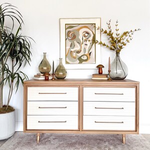 May include: A mid-century modern style dresser with six drawers. The drawers are white with horizontal grooves and brass handles. The dresser is made of light wood and has gold-colored legs. The top is decorated with vases, candles, and a framed abstract art piece.