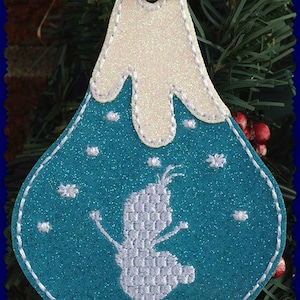 May include: A blue and white felt Christmas ornament with a snowman design. The ornament is shaped like a mountain with a white snowy peak. The snowman is white and has a simple design. The ornament is decorated with white snowflakes. The text "MMD Embroidery" is visible at the bottom of the ornament.