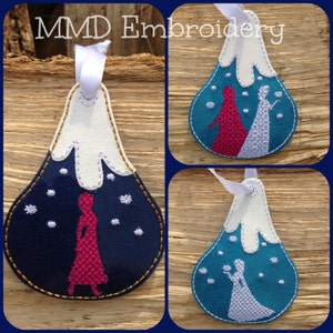 May include: Three felt ornaments with embroidered images of snow-covered mountains and figures in winter clothing. The ornaments are blue, teal, and white with gold stitching.