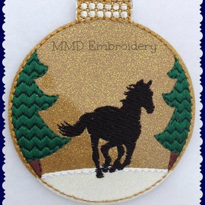May include: A gold glitter Christmas ornament with a black silhouette of a horse running through a snowy scene with two green pine trees on either side. The ornament has the text "MMD Embroidery" on it.