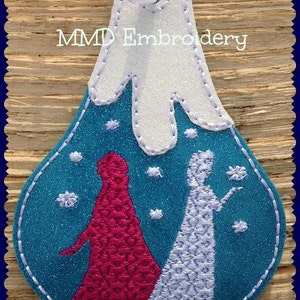 May include: A teal blue felt ornament with white felt snow and two embroidered figures of women in white and pink dresses. The ornament has a white felt mountain top and the text "MMD Embroidery" at the top.