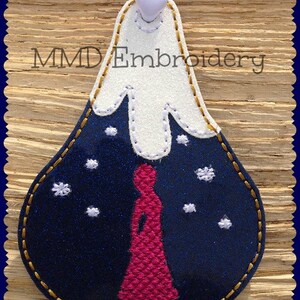 May include: A blue felt ornament with a white felt mountain top and a pink felt figure standing in front of it. The ornament is decorated with white snowflakes and has a gold stitched border. The text "MMD Embroidery" is visible at the top of the ornament.