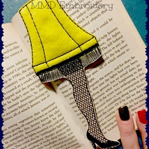 May include: A yellow felt bookmark shaped like a lampshade from the movie 'A Christmas Story'. The bookmark is attached to a black felt leg with a black fishnet stocking. The bookmark is placed on a book with text about a cemetery.