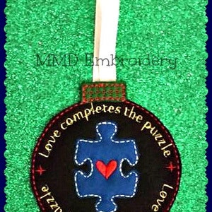 May include: A black and blue Christmas ornament with a puzzle piece design and the text "Love completes the puzzle" repeated twice. The puzzle piece has a red heart in the center.