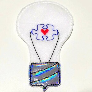 May include: White felt lightbulb with a blue puzzle piece with a red heart in the center. The lightbulb has a silver iridescent base.
