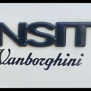 May include: A white van with a black "TRANSIT" emblem and a black cursive "Vanbozghini" sticker on the back.