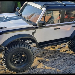 Puede incluir: Un modelo de SUV a control remoto de color blanco con detalles en negro, neumáticos todoterreno negros y llantas negras. El vehículo presenta un gráfico de silueta de caballo negro en el capó y el panel lateral, además de una baca negra.