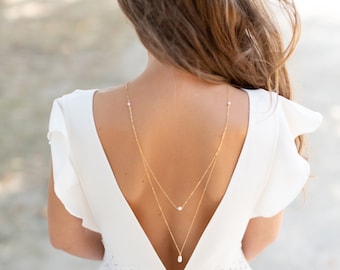 May include: A white bridal dress with a low back and a lace trim. The dress features a delicate gold chain with pearl accents.