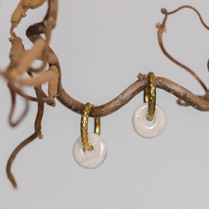 May include: A pair of gold-toned hoop earrings with a textured design, each holding a white, circular pendant with a marbled effect. The earrings are suspended from a brown, twig-like branch against a soft, neutral background.
