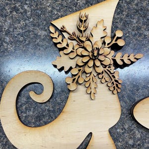 May include: Wooden Christmas elf shoe decoration with a decorative snowflake and leaf design. The elf shoe is made of light-colored wood and has a curved top and a cut-out design.
