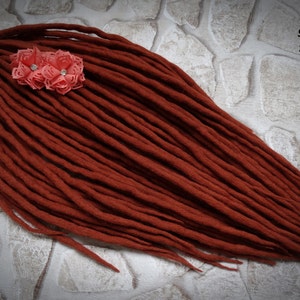 Puede incluir: Un conjunto de rastas largas de lana de color marrón rojizo con un acento de flor rosa.