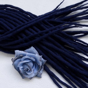 Puede incluir: Un conjunto de rastas azul marino oscuro, dispuestas sobre una superficie blanca, con una rosa de tela azul claro con detalles de purpurina plateada. Las rastas son gruesas y cilíndricas.