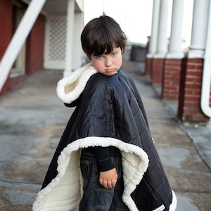 May include: A young person wearing a black hooded poncho with a white sherpa lining. The poncho has a quilted design and a white label with black text that reads "Sleepy Time Snuggles". The person is wearing blue jeans and grey sneakers.