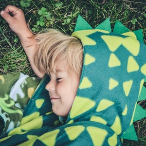 May include: A child wearing a green and yellow dinosaur costume with a hood lies on their back in a grassy area.