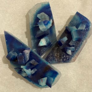 Puede incluir: Tres objetos con forma de cristal azul y blanco. Cada uno tiene un centro azul oscuro, desvaneciéndose hacia bordes azules claros y blancos. Pequeños trozos cristalinos azul claro están dispersos encima, creando una apariencia texturizada. Dispuestos sobre una superficie blanca.