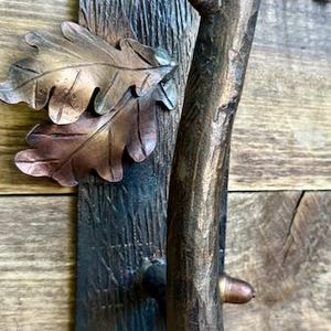 May include: A rustic bronze-toned door handle with a tree branch design. The handle features a textured branch grip, a decorative plate with oak leaves, and is mounted on a wooden surface. The handle is approximately 10 inches in height.