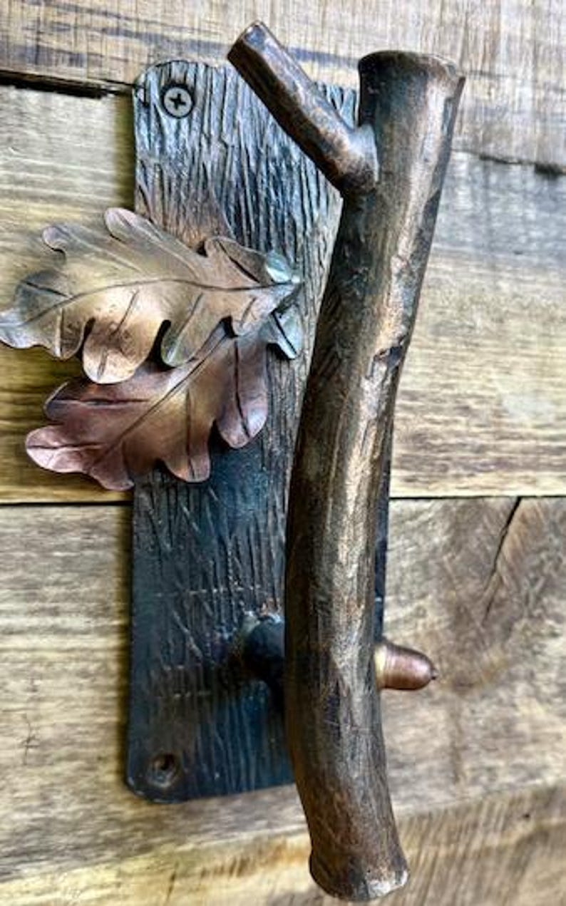 May include: A rustic bronze-toned door handle with a tree branch design. The handle features a textured branch grip, a decorative plate with oak leaves, and is mounted on a wooden surface. The handle is approximately 10 inches in height.