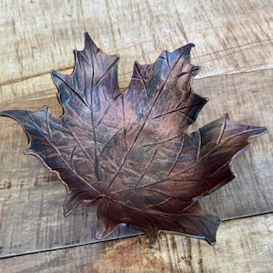 May include: A decorative leaf-shaped bowl with a bronze finish. The bowl is designed to resemble a maple leaf, with detailed veining and a textured surface. The bowl is made of metal and is suitable for holding small items.