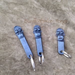 May include: Three silver keychains with black metal monkey faces. The keychains are on a brown textured surface. The text "Burnt Whisker Forge" is visible above the keychains.