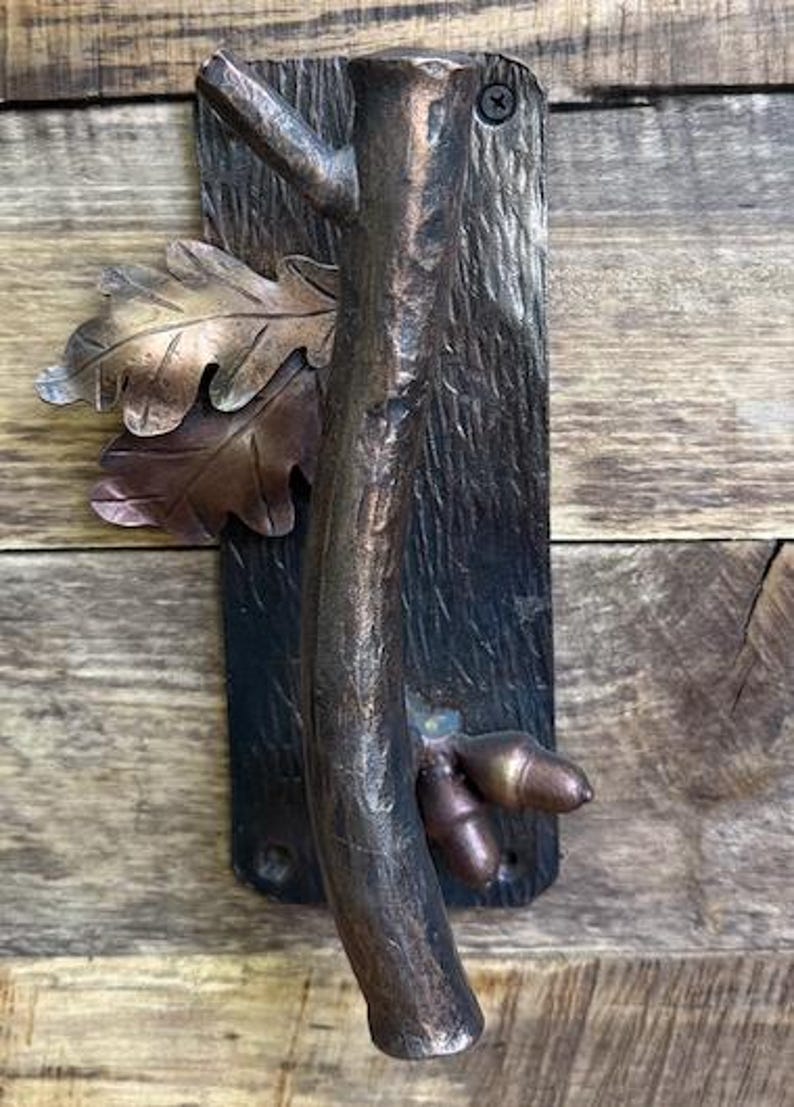 May include: A rustic bronze door handle with a tree branch design. The handle features a textured tree trunk, oak leaves, and acorns. The handle is mounted on a dark rectangular plate, perfect for a cabin or nature-themed decor.